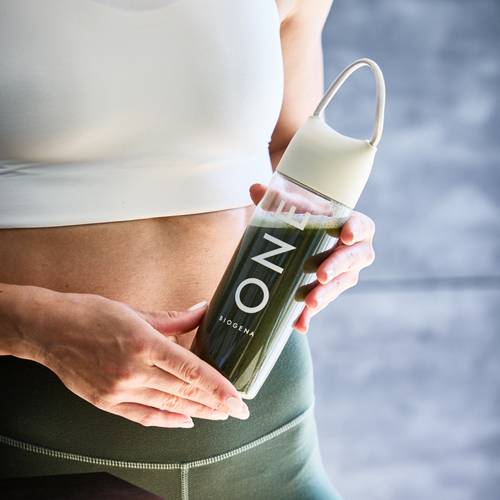 Person, die eine Flasche mit einem grünen Getränk hält, trägt ein weißes Sport-BH und dunkle Leggings vor einem unscharfen Hintergrund.