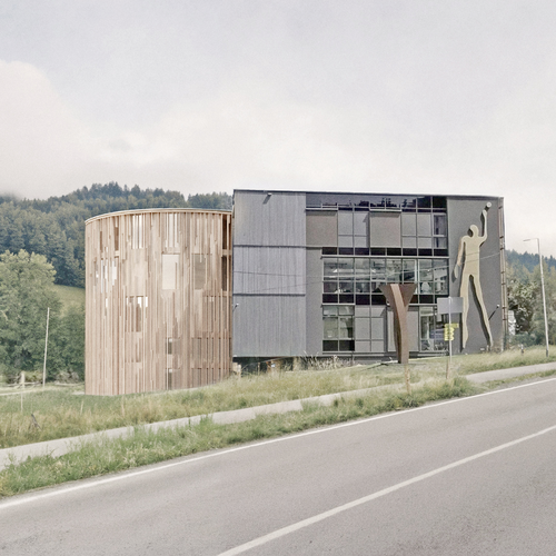 Ein modernes Gebäude mit einer runden Fassade aus Holz und Glas, an der ein kunstvolles Silhouettenbild zu sehen ist, vor dem Hintergrund von Bäumen und einer Straße.