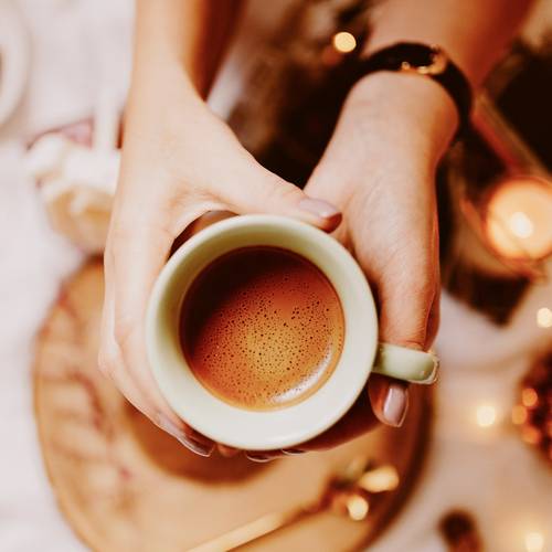 Mâini care țin o cană verde plină cu cafea, înconjurate de o atmosferă primitoare și lumini calde.
