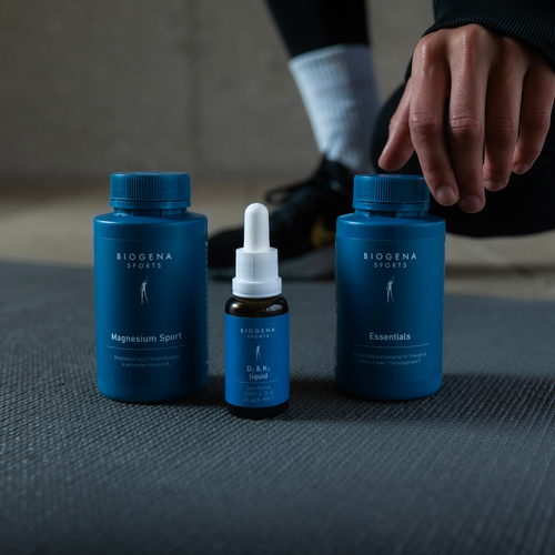 Trois compléments alimentaires BIOGENA SPORTS, dont Magnesium Sport, Essentials et D&K Liquid, sont posés sur un tapis, à côté duquel une personne est agenouillée.