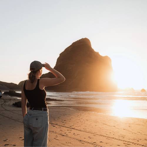 Eine Frau mit Hut steht am Strand bei Sonnenuntergang und blickt auf eine große Felsformation am Meer.