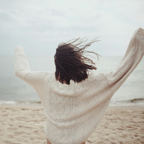 Una persona con un maglione morbido, in piedi sulla spiaggia con le braccia alzate, mentre il vento le scompiglia i capelli e lei guarda il mare sotto un cielo nuvoloso.