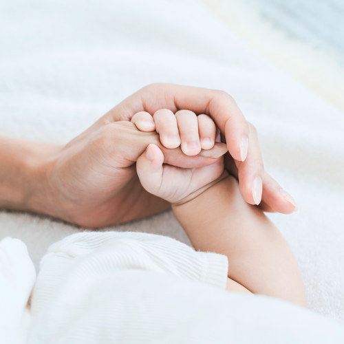 Die Hand eines Erwachsenen, die sanft die Hand eines Babys auf einer weichen, weißen Decke hält – ein Symbol für Fürsorge und Schutz.