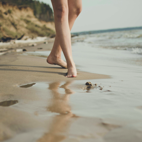 Une personne marche pieds nus sur une plage de sable, tandis que de douces vagues viennent lécher le rivage et qu'on aperçoit, en arrière-plan, une falaise boisée.