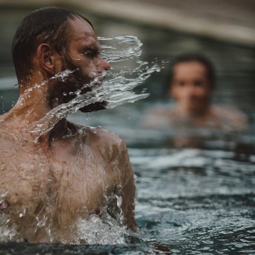 Ein Mann im Schwimmbecken dreht den Kopf und spritzt dabei Wasser in die Luft, während im Hintergrund eine weitere Person zu sehen ist.