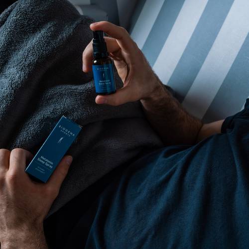 Person mit einer blauen Flasche und einer Schachtel mit der Aufschrift „Melatonin Sleep Spray“ auf einem gestreiften Bett, teilweise bedeckt von einer grauen Decke.