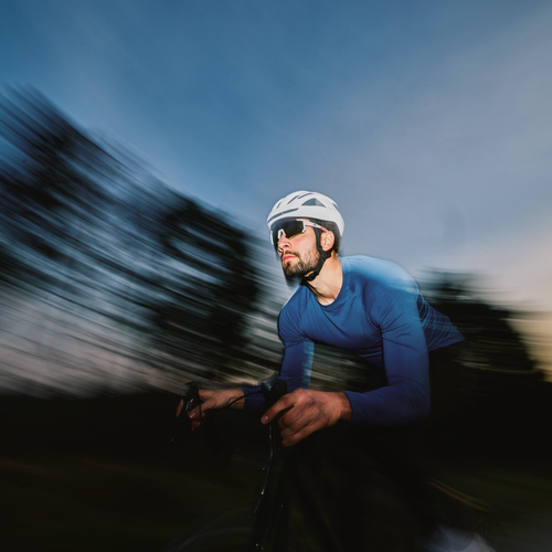 Un ciclista con camiseta azul y casco blanco recorre rápidamente un paisaje borroso al atardecer, creando una sensación de movimiento y velocidad.