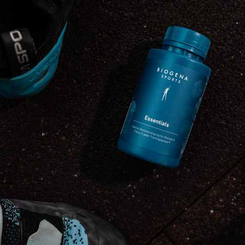 Sticlă albastră Biogena Sports Essentials pe o suprafață întunecată și texturată, cu părți din două pantofi vizibile în apropiere.