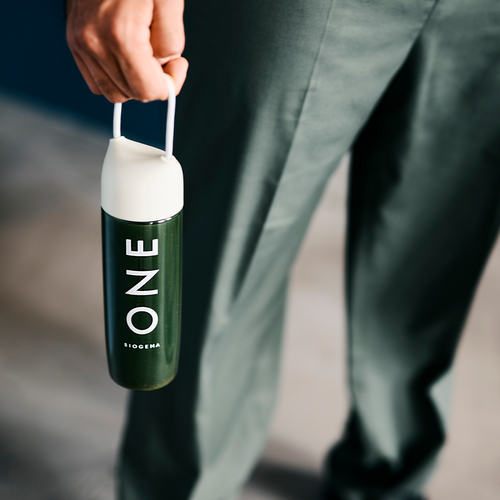 Persoană ținând o sticlă verde cu eticheta BIOGENA ONE cu capac alb și buclă, purtând pantaloni verde închis.