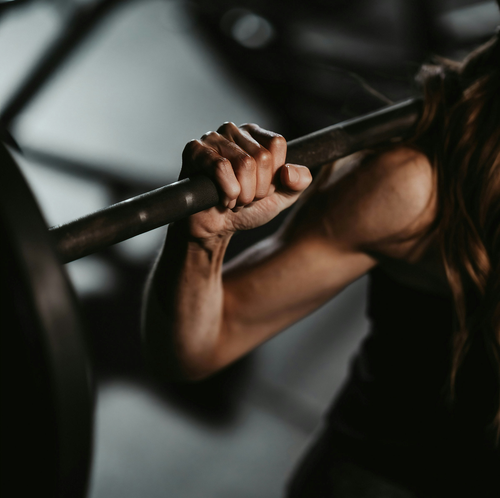 Nahaufnahme einer Person, die eine Hantel stemmt, wobei der Fokus auf der Hand liegt, die die Stange umklammert, und die muskulöse Armkraft in einem schwach beleuchteten Fitnessstudio zur Geltung kommt.