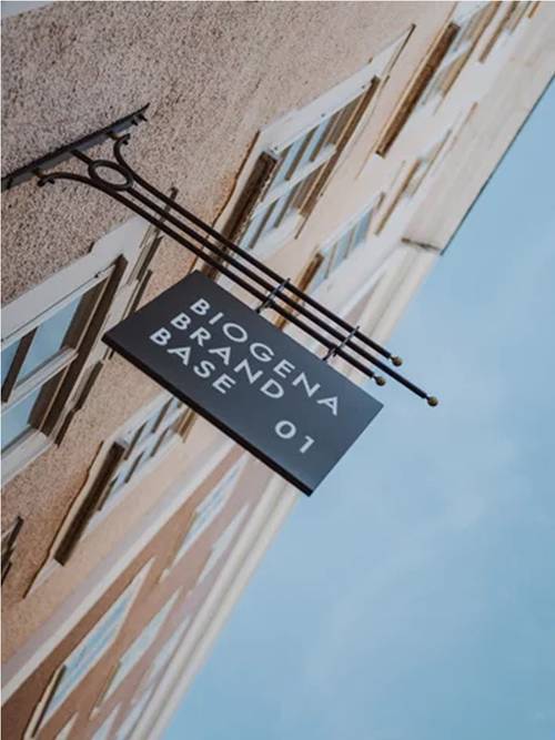 Schild mit der Aufschrift "BIOGENA BRAND BASE 01" an der Wand eines Gebäudes, von unten gesehen gegen einen blauen Himmel.
