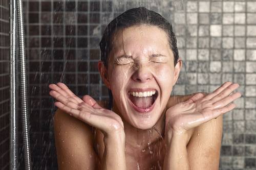 Person in a shower with eyes closed, beaming smile and hands in front of face. Water flowing down a tiled wall.