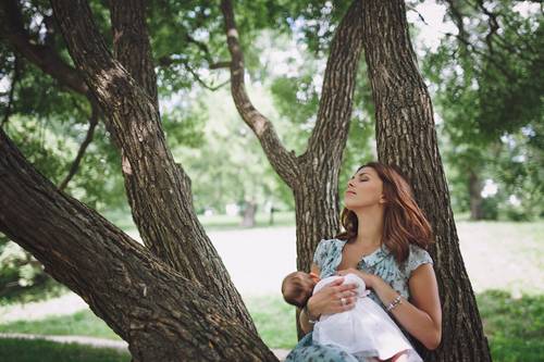 A woman sits against a tree with her eyes closed, peacefully holding a baby in a park setting.
