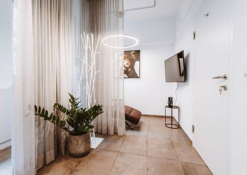 Modernes Zimmer mit einer Topfpflanze, transparenten Vorhängen, einer einzigartigen Stehlampe, einem an der Wand montierten Fernseher und Kunstwerken. Neutrale Farbtöne sorgen für eine minimalistische Atmosphäre.