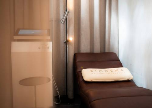Gemütlicher Wellnessraum mit Liegestuhl, „Biogena”-Kissen, sanfter Beleuchtung und einem medizinischen Gerät, das teilweise hinter einem transparenten Vorhang zu sehen ist.