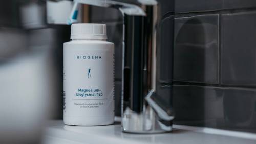 Eine Flasche BIOGENA Magnesium-Bisglycinat 125, die neben einem Metallwasserhahn auf einem gefliesten Waschtisch im Badezimmer steht.