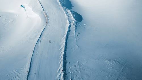 Luftaufnahme eines einsamen Skifahrers auf einem schneebedeckten Berghang unter strahlend blauem Himmel, mit sanften Schatten und strukturiertem Schnee.