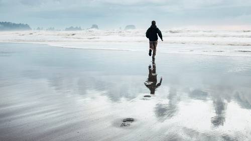 Eine Person rennt an einem nassen Sandstrand entlang, im Hintergrund sind Meereswellen und ein bewölkter Himmel zu sehen. Ihr Spiegelbild ist im Sand zu erkennen.