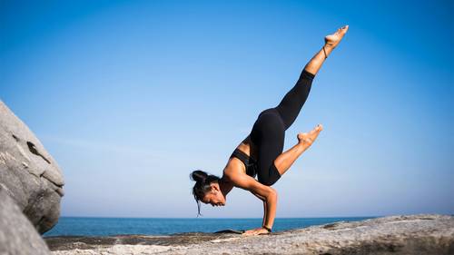O persoană face un stand pe mâini de yoga pe o suprafață stâncoasă lângă mare, sub un cer senin și albastru, purtând echipament sportiv negru.