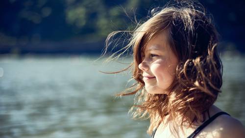 Chica con el cabello movido por el viento sonríe junto al agua, con un fondo natural desenfocado.