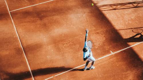 Un jucător de tenis îmbrăcat în albastru servește pe un teren de zgură, proiectând o umbră, cu mingea verde estompată în aer.