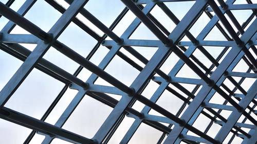 Prim-plan al unei structuri geometrice din grilaj metalic pe un fundal deschis, creând un model de linii și unghiuri care se intersectează.
