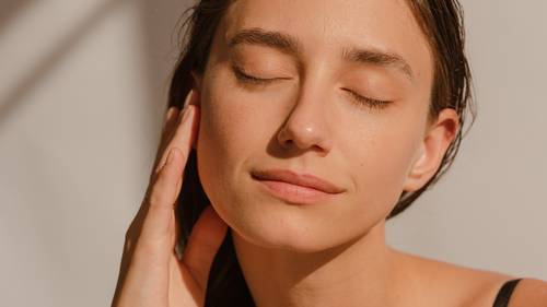A woman with closed eyes gently touches her face, enjoying the warmth of sunlight on her skin, with a serene expression.