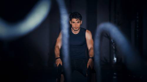 Bărbat antrenându-se intens cu frânghii de luptă într-o sală de sport întunecată, purtând un maiou negru
