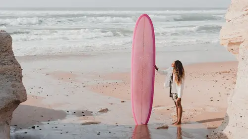 A person stands on a sandy beach holding a pink surfboard, with waves crashing in the background and rocky cliffs on either side.