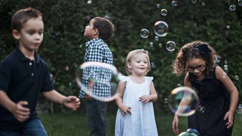 Vier Kinder spielen fröhlich mit Seifenblasen in einem Garten. Sie lächeln und tragen legere Sommerkleidung, umgeben von grünen Pflanzen.