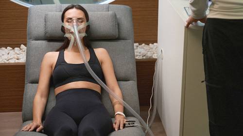 Eine Frau in Sportkleidung sitzt auf einem Liegestuhl und trägt eine Sauerstoffmaske, während eine weitere Person daneben steht.