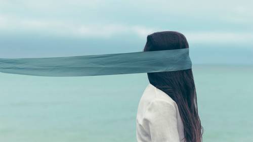 A woman with long hair stands by the sea, her eyes covered by a flowing blue scarf, looking towards the horizon under a cloudy sky.