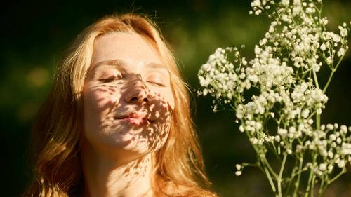 Eine Frau mit geschlossenen Augen genießt das Sonnenlicht, das die Schatten weißer Blumen auf ihr Gesicht wirft, umgeben von Grünpflanzen.