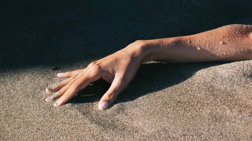Eine Hand, die teilweise im Sand vergraben ist und deren Finger gespreizt sind, wirft einen Schatten auf die raue Oberfläche.