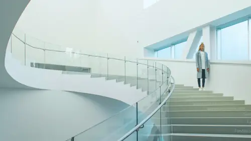 A person stands on a modern, curved staircase with glass railings in a bright, minimalist interior.