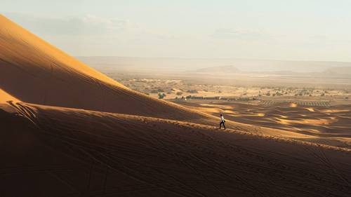 A person walks on a vast, sunlit sand dune with a distant landscape of smaller dunes and a hazy sky in the background.