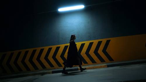 Silhouetted person walking in a dimly lit area with a yellow and black chevron pattern on the wall and a bright overhead light.