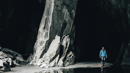 Eine Person in einer blauen Jacke erkundet eine große, dunkle Höhle mit hoch aufragenden Felsformationen und verstreuten Steinen auf dem Boden.