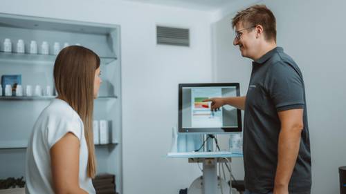 Eine Person in einem dunklen Shirt zeigt auf einen Computerbildschirm, während eine andere Person in einem modernen Büro aufmerksam zuhört.