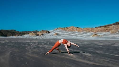 Persoană îmbrăcată în echipament sportiv portocaliu execută o postură de yoga pe o plajă vastă cu nisip, cu dealuri stâncoase și cer senin în fundal.