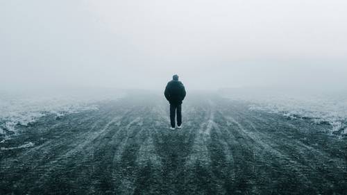 Eine Person geht allein auf einer nebligen, verlassenen Straße, umgeben von Nebel und kahlem, frostigem Boden, was ein Gefühl von Einsamkeit und Geheimnis erzeugt.