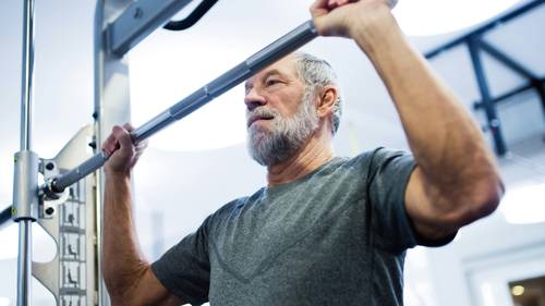 Ein älterer Mann mit Bart, der in einem Fitnessstudio eine Hantel stemmt; er trägt ein graues T-Shirt und ist ganz auf seine Übung konzentriert.