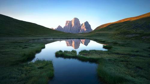 Eine friedliche Berglandschaft mit einem ruhigen See, der schroffe Gipfel spiegelt, umgeben von saftigem grünen Gras unter einem klaren blauen Himmel.