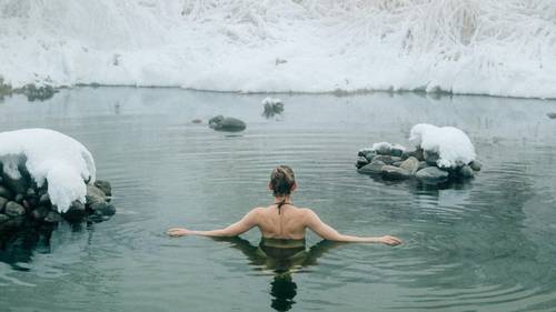 Person in einem Badeanzug, die in einer ruhigen heißen Quelle badet, umgeben von schneebedeckten Felsen und winterlicher Landschaft.