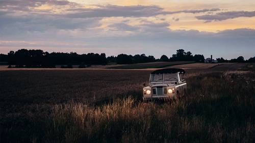 Klassisches Geländefahrzeug mit eingeschaltetem Scheinwerfer, das bei Dämmerung durch ein grasbewachsenes Feld unter wolkenverhangenem Himmel fährt.