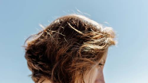 Close-up of a person with tousled brown hair against a clear blue sky, capturing the texture and movement of the hair.