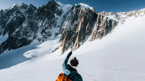 Eine Person in Winterkleidung zeigt auf schneebedeckte Berggipfel unter klarem blauen Himmel.