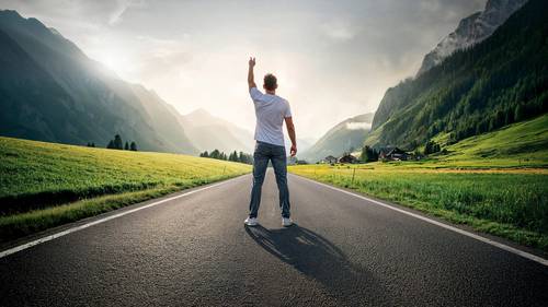 Eine Person steht auf einer leeren Straße, hebt eine Hand und ist von üppig grünen Bergen unter einem bewölkten Himmel umgeben.