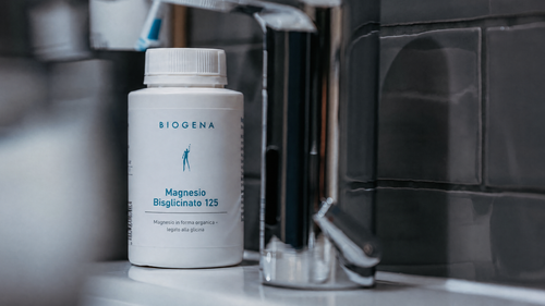 Una bottiglia di BIOGENA Bisglicinato di magnesio 125, appoggiata su un lavabo piastrellato in bagno, accanto a un rubinetto in metallo.
