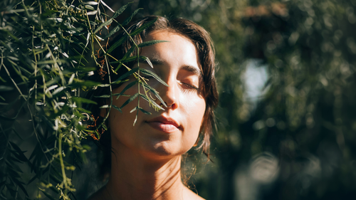 Woman with closed eyes, surrounded by sunlit green leaves, appearing tranquil.